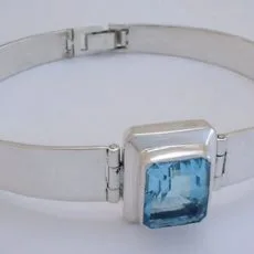 Front perspective view of a handcrafted 925 sterling silver hinged bangle bracelet featuring a sleek polished flat band and a central emerald-cut blue topaz gemstone in a raised bezel frame.