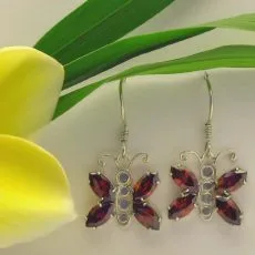 Pair of handcrafted 925 sterling silver butterfly drop earrings featuring marquise-cut deep red garnet wings, delicate wirework antennae, and french wire ear hooks resting next to a yellow frangipani flower.