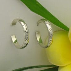 A pair of polished 925 sterling silver J-hoop earrings featuring intricate hand-engraved dot patterns, displayed on a smooth light surface next to a bright yellow frangipani flower.
