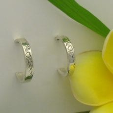 A pair of polished 925 sterling silver J-hoop earrings featuring hand-engraved heart and botanical patterns, displayed on a light background next to a yellow frangipani flower.