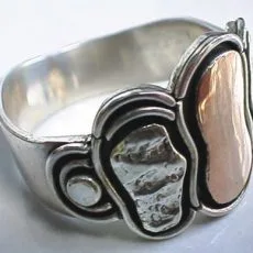 A close-up three-quarter view of an artisan-crafted wide 925 sterling silver statement ring showcasing an abstract organic design with a textured silver pebble and a contrasting smooth brass pebble inlay.