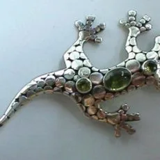 An artisan-crafted 925 sterling silver gecko brooch featuring a textured pebble-style body accented by three bezel-set green peridot gemstones and an oxidized finish.