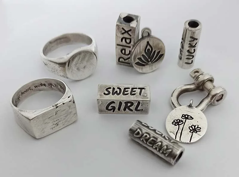 A flat-lay view showcasing a collection of custom engraved 925 sterling silver jewelry components, including textured signet rings, a shackle clasp with a floral disk, and oxidized rectangle and cylinder beads stamped with inspirational text.